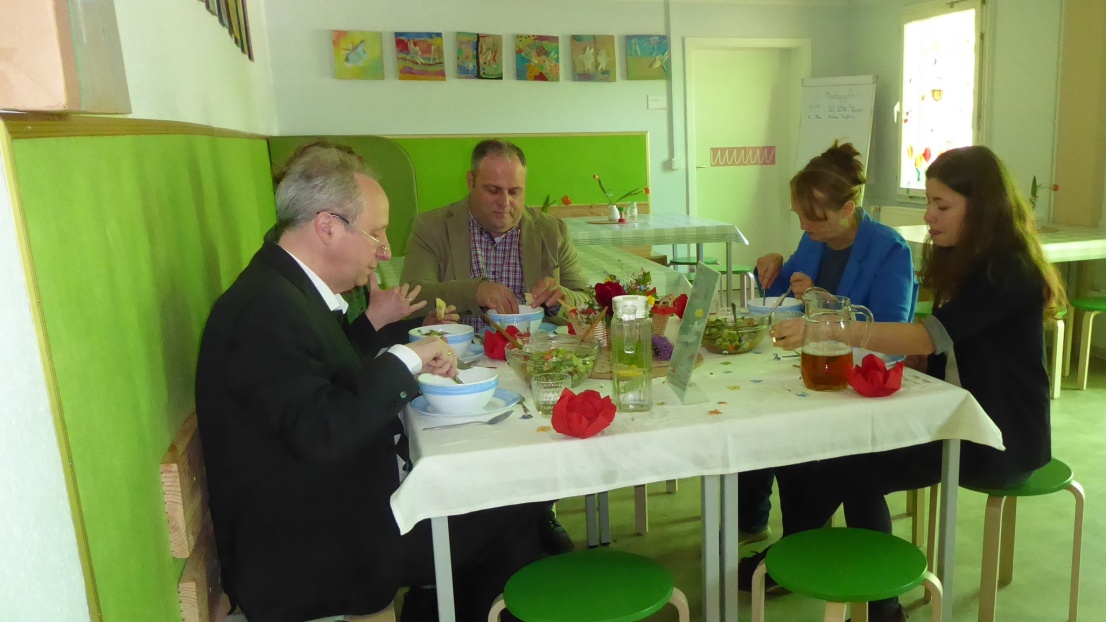 5 Menschen sitzen an einem bunt gedeckten Frühstückstisch und unterhalten sich, während sie essen.