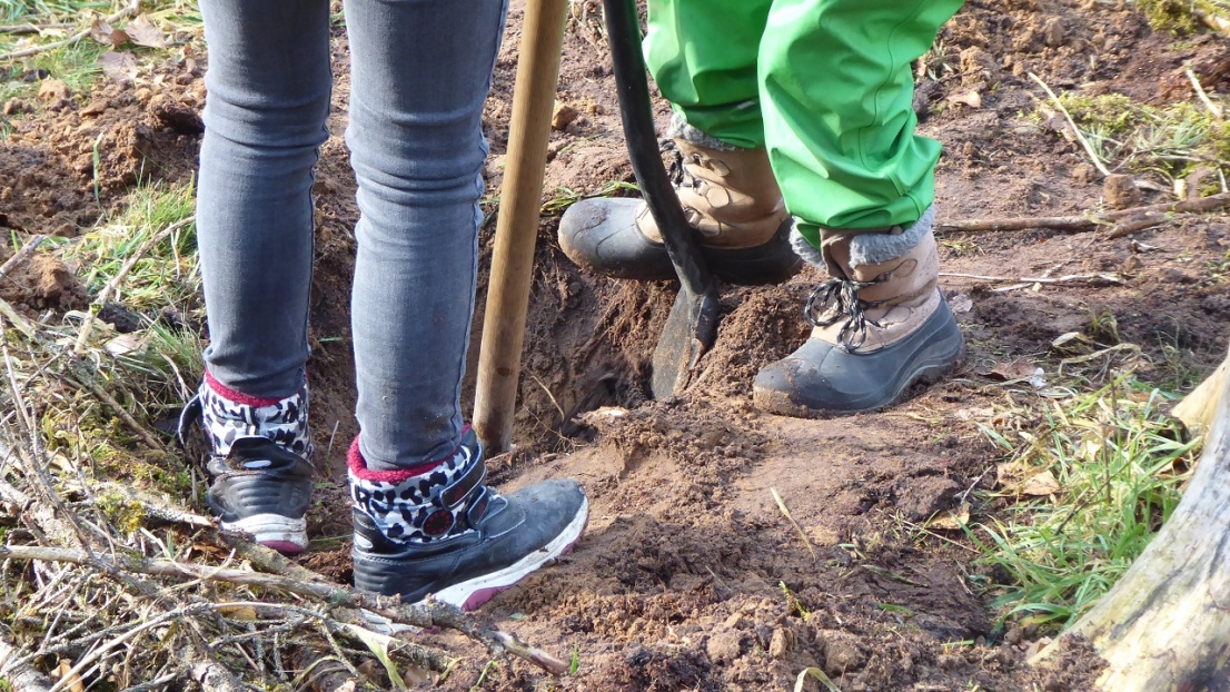 Man sieht die Beine von zwei Kindern, die mit Stiefeln und Matschhose ein Loch in der Erde graben