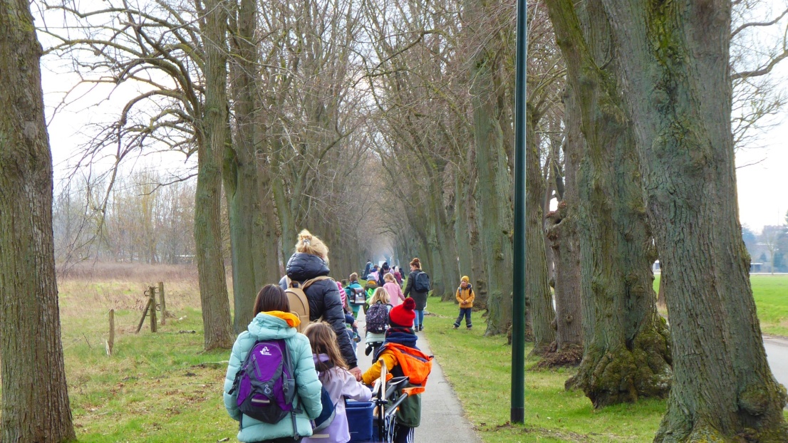 Auf einem ländlichen Fußgänger-Weg laufen bunt und für kühles Wetter gekleidete Kinder und Erwachsene. Sie tragen Schaufeln und andere Geräte im Gepäck.