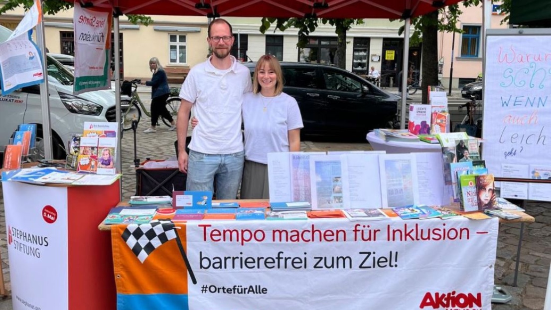 Ein Mann und und eine Frau stehen lächelnd Arm in Arm hinter einem Aktionsstand der Stephanus-Stiftung auf einem Marktplatz. Der Stand ist bunt und hält viele Informationsblätter und Plakate zum Thema Leichte Sprache bereit.