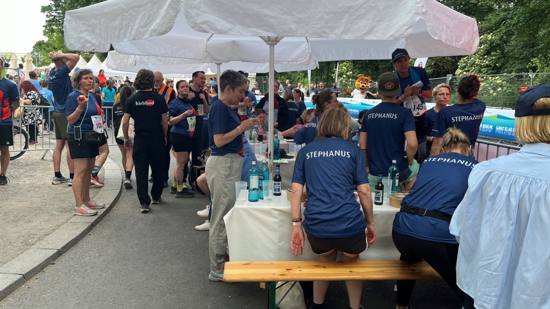 Eine Gruppe von fröhlichen Menschen steht und sitzt bei warmem Wetter unter weißen Sonnenschirmen. Sie tragen alle das gleiche Trikot mit dem Stephanus-Glockenstuhl-Logo. Auf den Biertischen stehen viele Getränke.