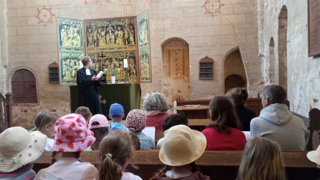 Mehrere Erwachsene und Kinder sind vom Rücken aus sichtbar: Sie sitzen in einer Kirche und schauen in Richtung Altar, wo eine Pfarrerin spricht. Sie sind sommerlich bekleidet.