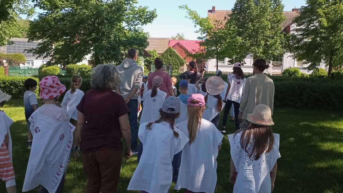 Mehrere Erwachsene und Kinder sind vom Rücken aus sichtbar: Sie stehen auf einer Wiese und tragen weiße Gewänder, die mit der Jakobsmuschel teilweise bestempelt sind