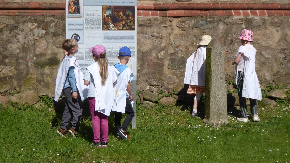 5 Kinder spielen auf einer Wiese neben einem alten Steingemäuer einer Kirche. Sie tragen weiße Gewänder und Sonnenhüte.