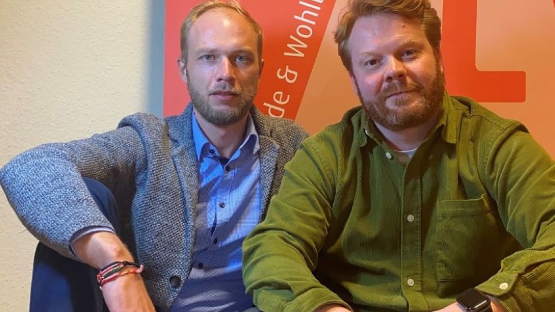 Zwei Männer sitzen locker im Schneidsitz zusammen und lächeln in die Kamera, einer davon ist der neue Bereichsleiter Catering, Christian Jebauer