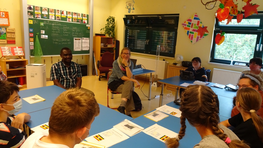 Ronald Mulondo sitzt in einem Klassenraum. An den Tischen sitzen Schüler*innen und Schüler. 