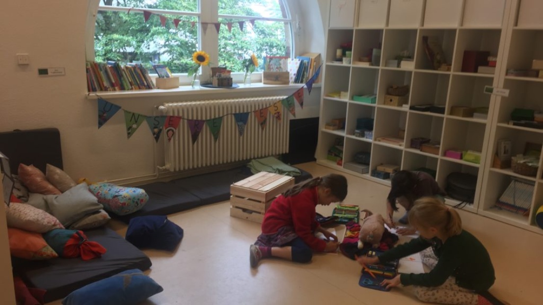 Eine gemütliche Leseecke mit Bücherregalen und Sitzkissen. Auf dem Boden sitzen Kinder und malen