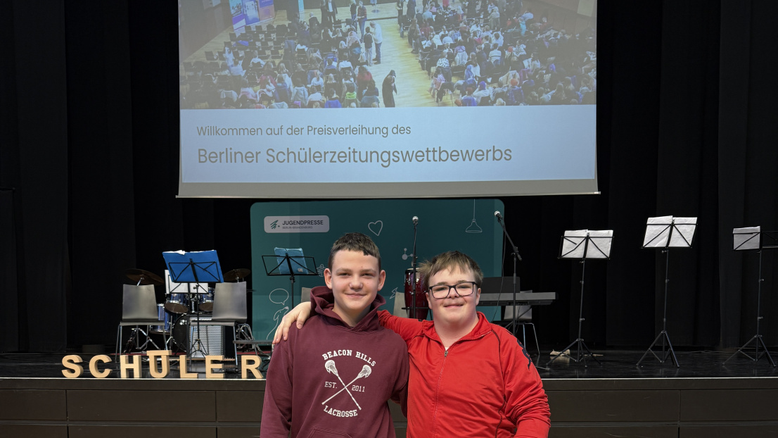 Zwei Jungs stehen zusammen vor einer Bühne. Auf der Leinwand hinten steht Schülerzeitungwettbewerb.