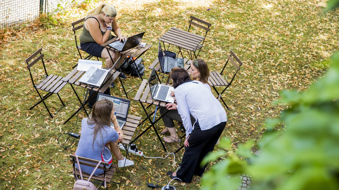 Drei junge Frauen sitzen im Garten mit ihren Laptops. Eine Frau beigt sich zu einer und schaut zu.