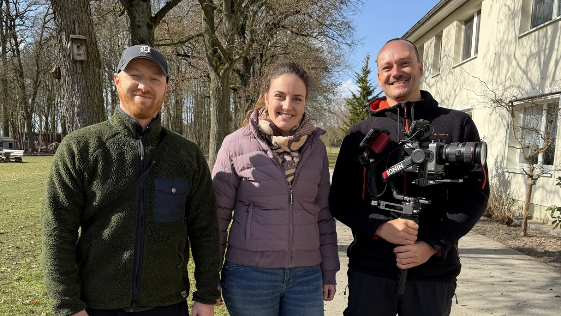 Drei Menschen stehen beieinander, lächeln in die Kamera, ein Mann hält eine Profi-Videokamera