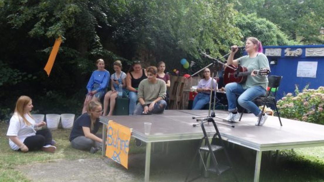 Eine junge Frau mit Gitarre sitzt auf der Bühne von Oberlin-Seminar. Um Sie herum sind Jugendliche.