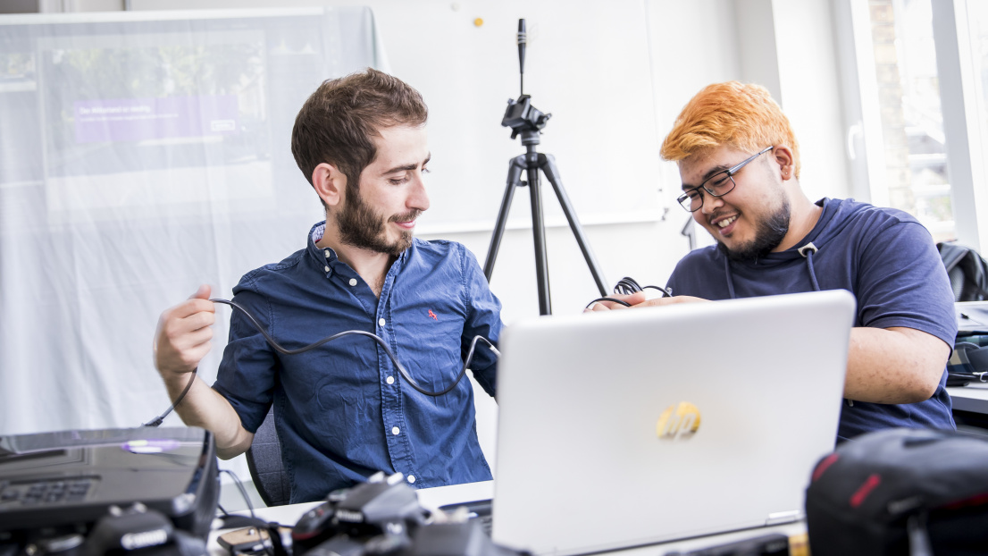 Zwei junge Männer sitzen am Laptop, Kameras liegen verteilt und sichtbar im ganzen Raum.