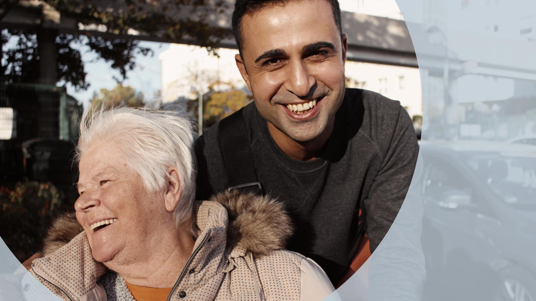 Ein junger Mann steht hinter einer Seniorin. Beide lachen. Der Mann schaut in die Kamera.