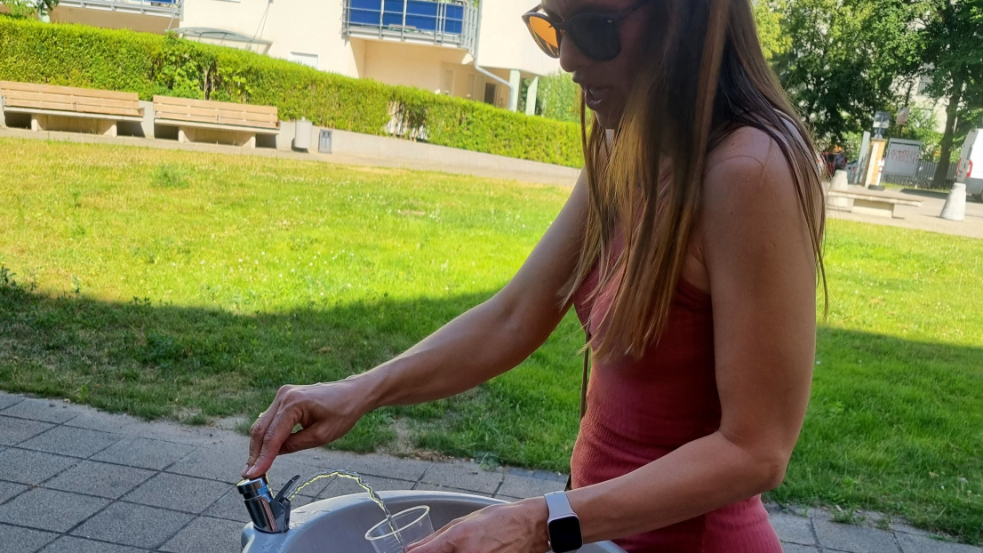 Frau steht an einem Trinkwasserspender und füllt Wasser in einen Becher.