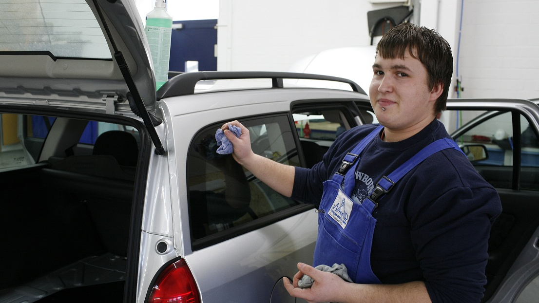 Ein junger Mann in einer Werkstatt macht ein Auto sauber und schaut lächelnd in die Kamera.