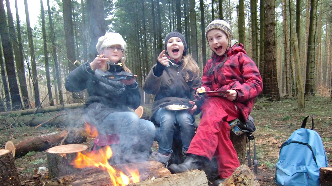 Drei Mädchen sitzen auf einem Baumstamm. Sie essen am Lagerfeuer und haben Spaß dabei.