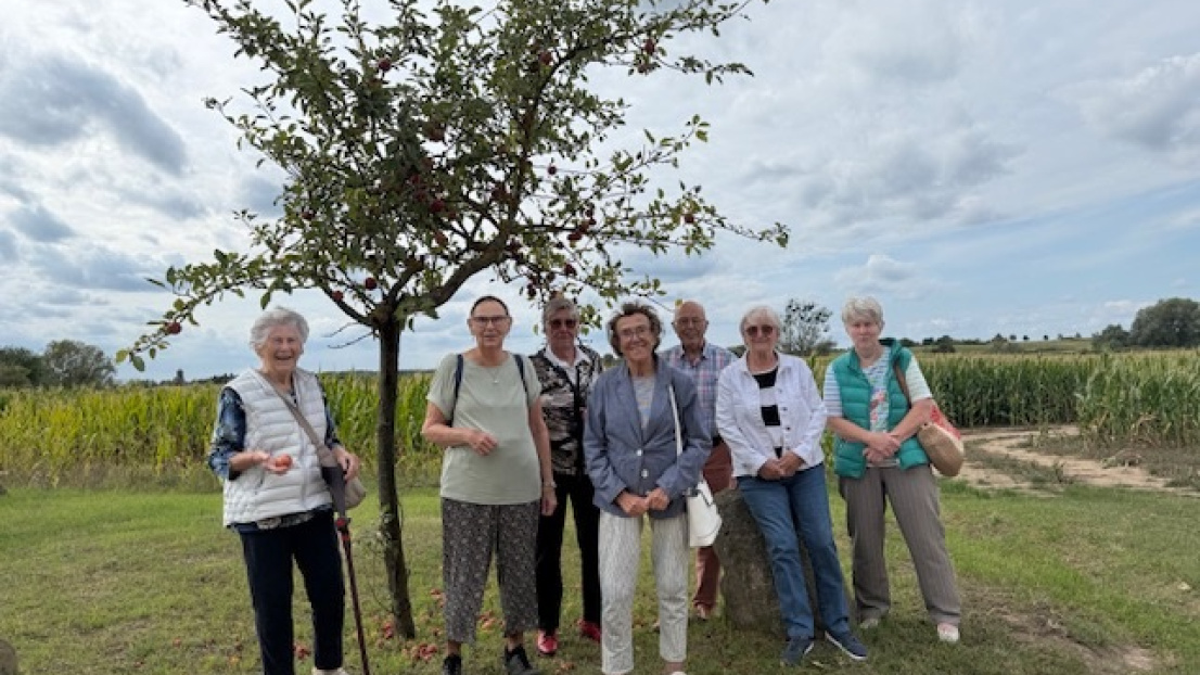 Sieben ältere fröhlich gelaunte Menschen stehen am Feld vor einem Baum.