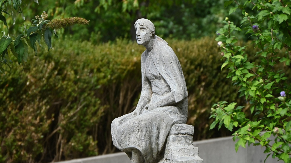 Skulptur einer sitzenden Frau auf Steinen. An der Skulptur liegen Rosen.