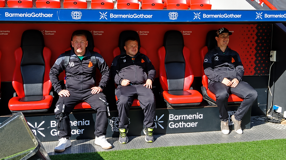 Drei Männer in Sportbekleidung mit dem Stephanus Glockenstuhl sitzen auf der Trainerbank.