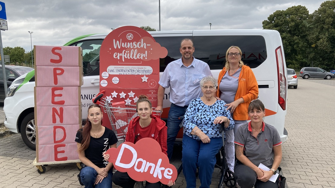 Eine Seniorin und sechs weitere Menschen stehen am Waagen mit Wunscherfüller-Spenden von Rossmann