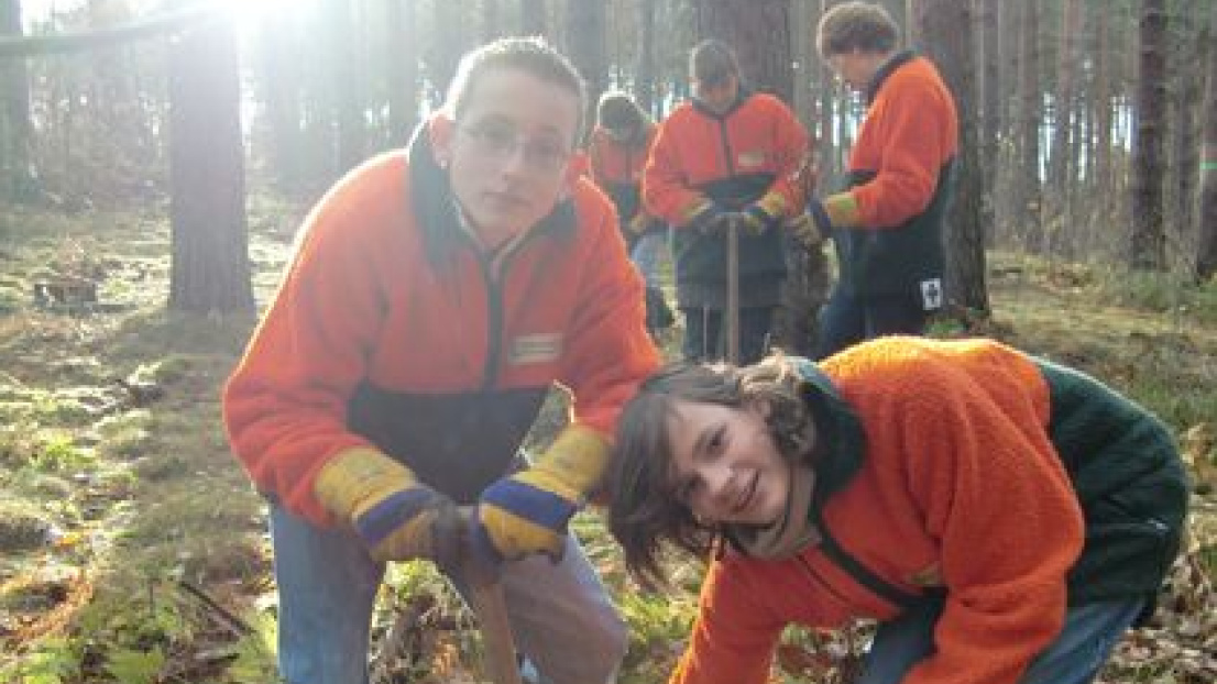 Jugendliche pflanzen in Wald. Alle haben orange-schwarzfarbigen Fleecejacken und Gummistiefel an.