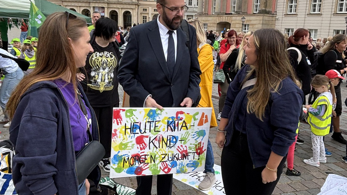 Zwei Frauen sprechen mit einem Mann in Anzug, der das handgemalte Plakat in den Händen hält.