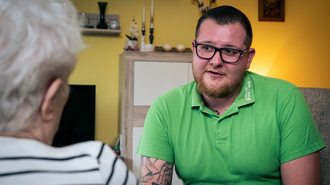 Ein Mitarbeiter im grünen Shirt im Gespräch mit einer Seniorin