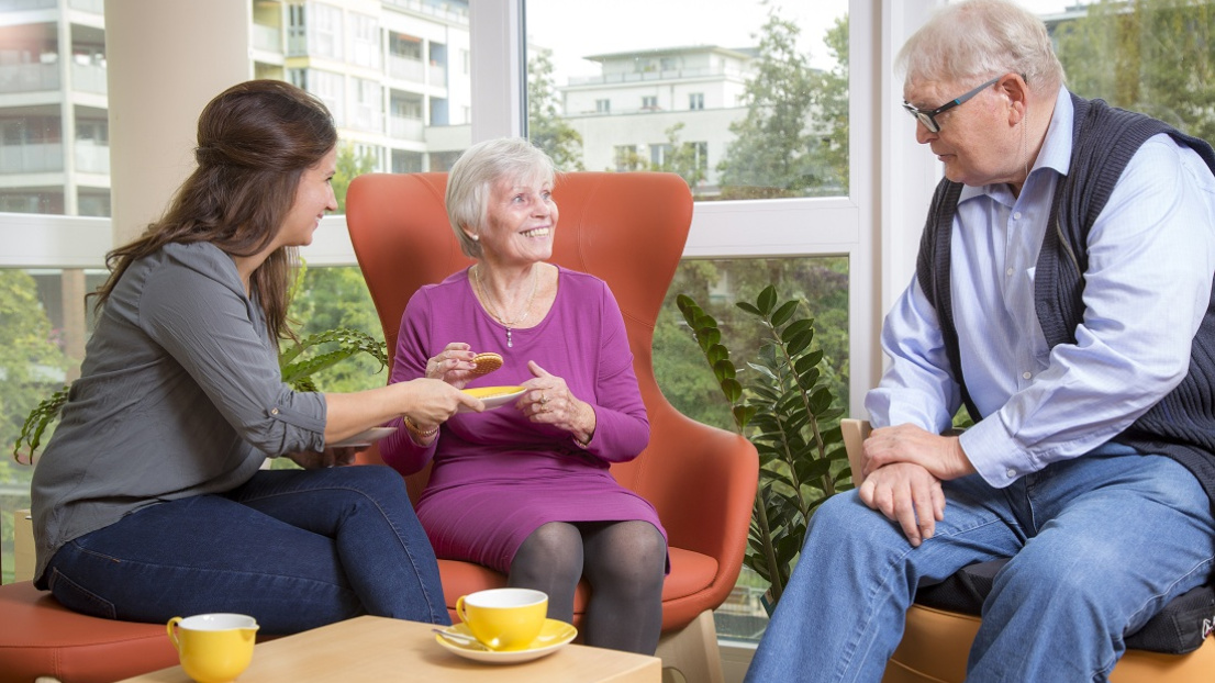 Eine Seniorin sitzt mit zwei Personen zusammen und genießt fröhlich Tee und einen Keks.