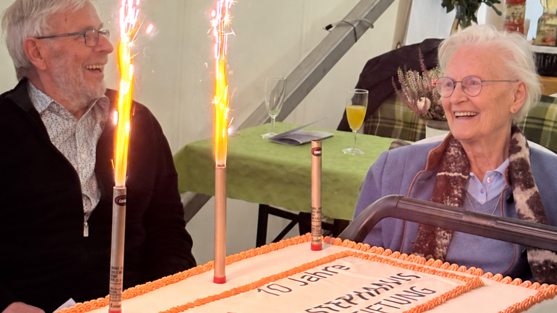 Eine Seniorin und ein Senior stehen an der Jubiläumstorte und lachen herzlich