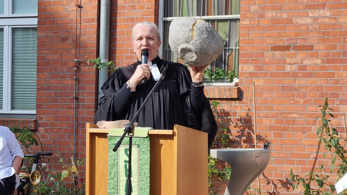 Pastorin steht am Altar, hält ein Mikrofon und eine Keramikschüssel.