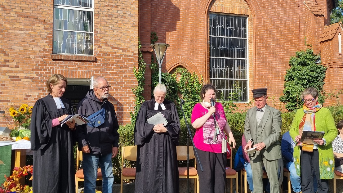 Zwei Pastorinnen und vier weitere Menschen stehen vor der Kirche. Eine Frau hält ein Mikrofon.