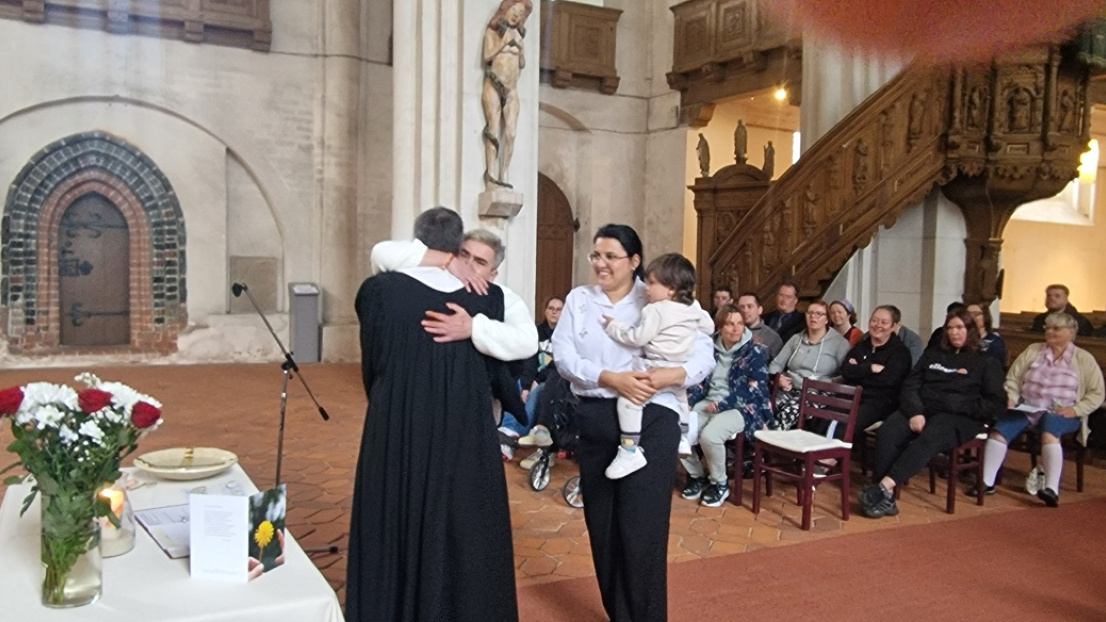 Ein Pastor umarmt eine Frau in der Kirche, während daneben eine junge Frau ein Baby auf dem Arm hält.