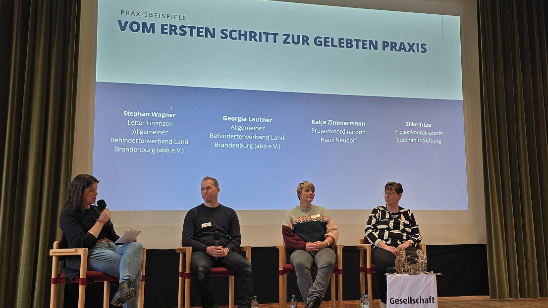 Vier Menschen sitzen auf einer Bühne mit einer Leinwand von Praxisbeispiele und Namen der Teilnehmer