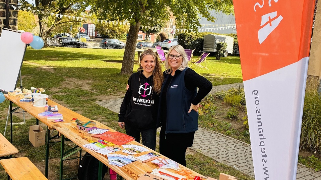 Zwei Frauen stehen am Tisch mit Flyer und Stephanus Werbeartikeln und lächeln in die Kamera.