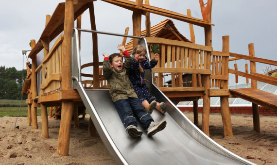 Zwei Kinder rutschen nebeneinander eine Rutsche von einem Spielturm herunter. 