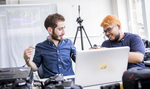 Zwei junge Männer sitzen am Laptop, Kameras liegen verteilt und sichtbar im ganzen Raum.