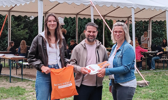 Auf dem Bild zu sehen: Celina Wöller und Stephanie Thiedig, Projektkoordinatorinnen, wie sie Michael Heber einen Beutel mit Spielzeug und etwas zu Naschen überreichen, im Hintergrund das Zelt mit Teilnehmenden am Kreativprogramm