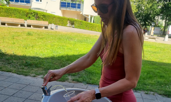 Frau steht an einem Trinkwasserspender und füllt Wasser in einen Becher.