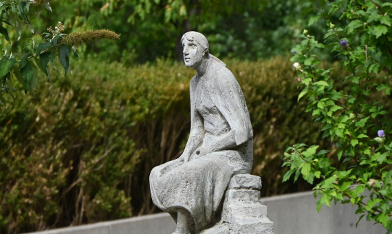 Skulptur einer sitzenden Frau auf Steinen. An der Skulptur liegen Rosen.