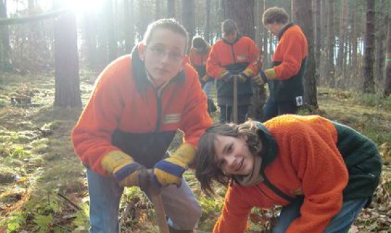 Jugendliche pflanzen in Wald. Alle haben orange-schwarzfarbigen Fleecejacken und Gummistiefel an.