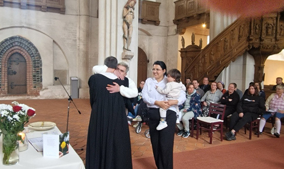 Ein Pastor umarmt eine Frau in der Kirche, während daneben eine junge Frau ein Baby auf dem Arm hält.