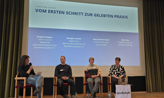 Vier Menschen sitzen auf einer Bühne mit einer Leinwand von Praxisbeispiele und Namen der Teilnehmer