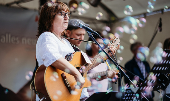 Person spielt Gitarre und singt auf einer Bühne, Seifenblasen fliegen durch die Luft.